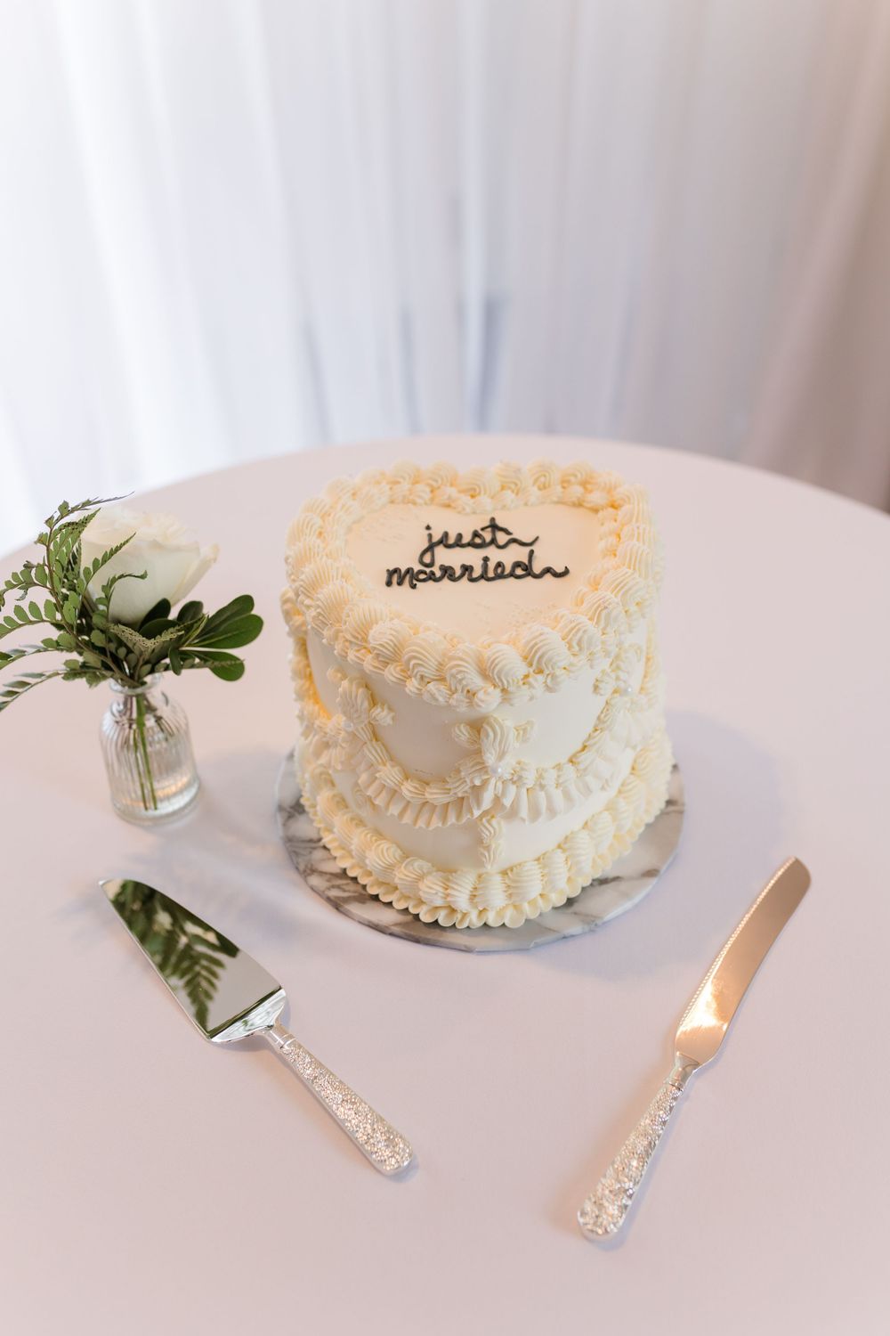 Two-tier white heart-shaped wedding cake with 'just married' topper, decorative piping, beside cake knife and server.