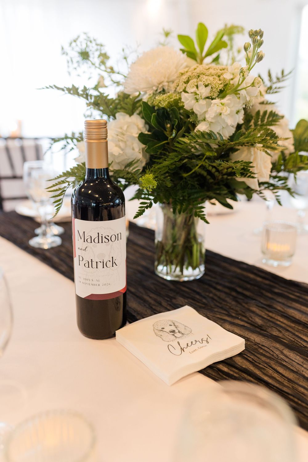 Custom Madison & Patrick wedding wine bottle on elegant table with white floral centerpiece and Cheers napkin.