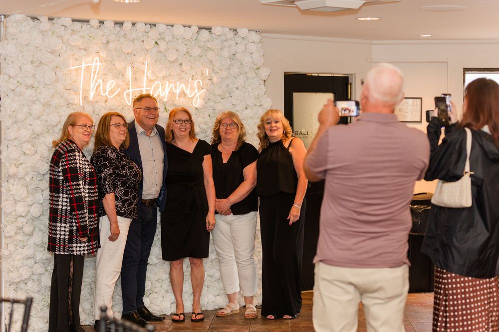 Group of people posing for a photo in front of a white floral wall with a neon sign reading The Harriss.