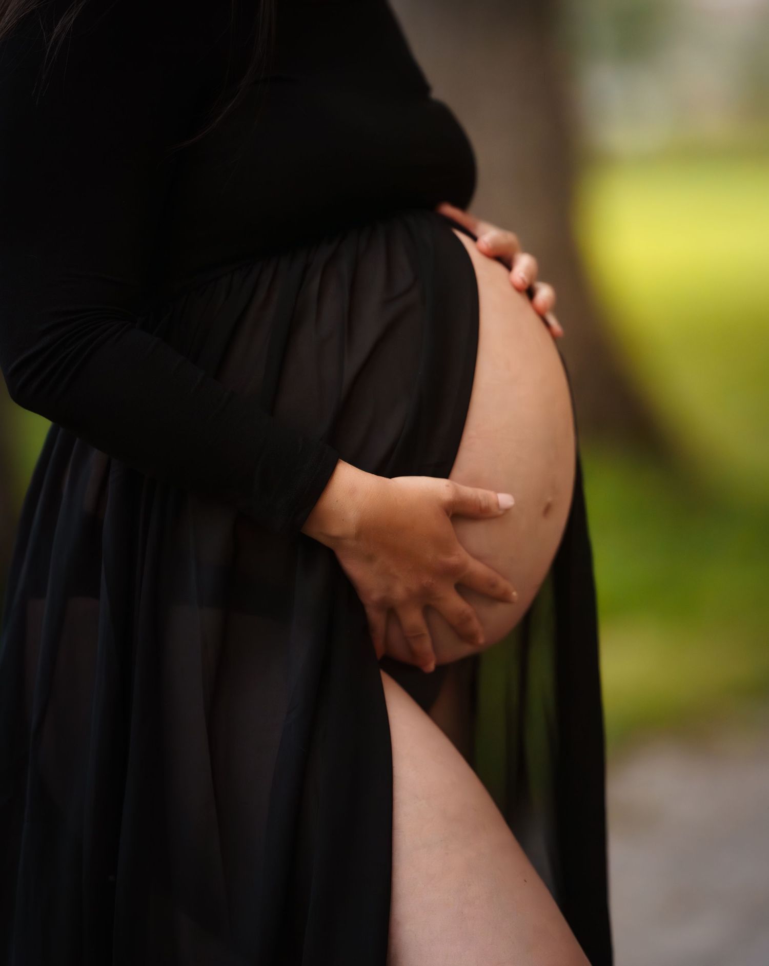Gros plan d'une future maman en robe noire qui tient son ventre arrondi à deux mains, lors d'une séance photo maternité en nature à Québec