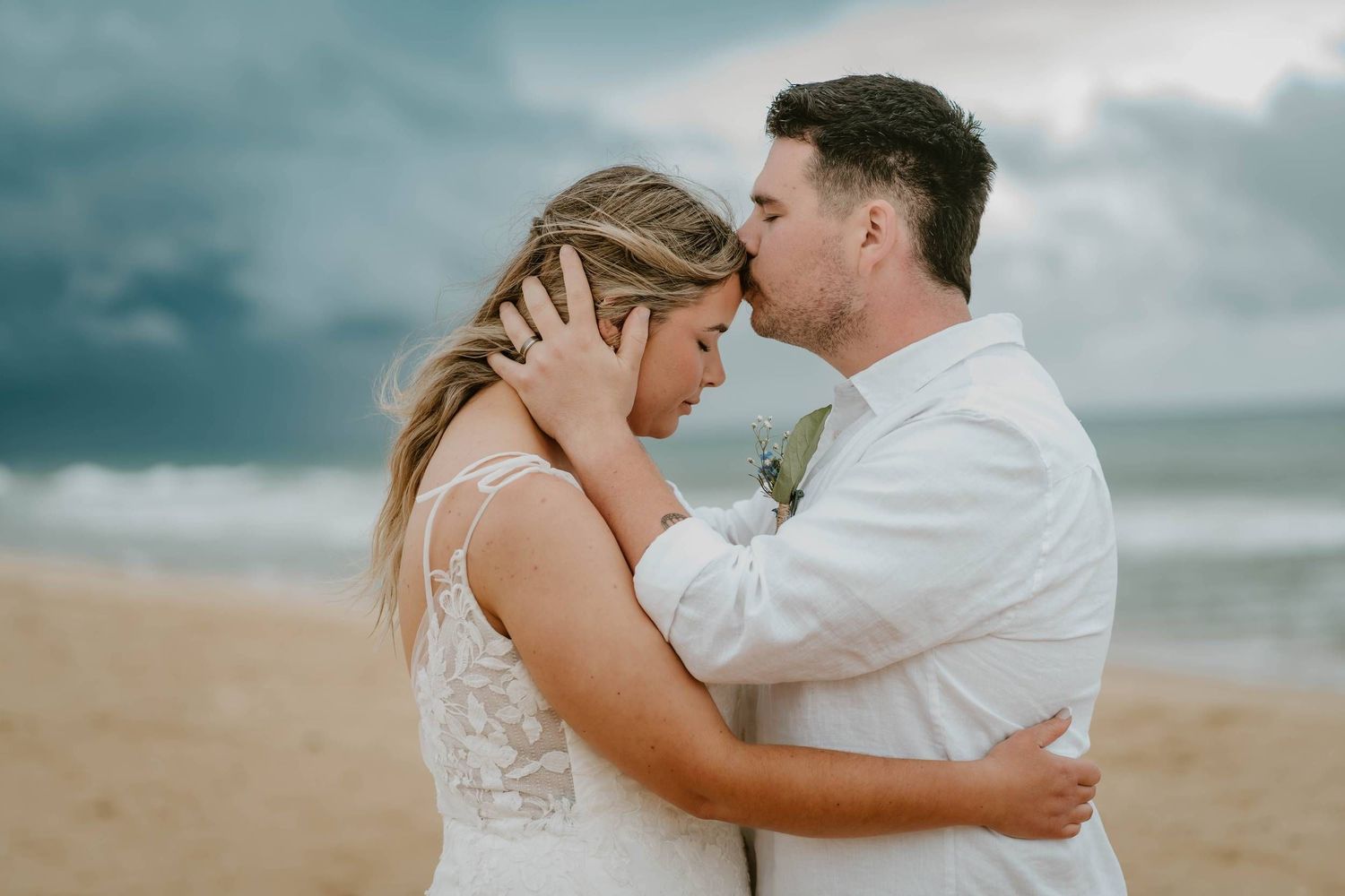 Bride and groom share a tender kiss on a beach with dramatic stormy skies, waves, and sandy shores behind them.