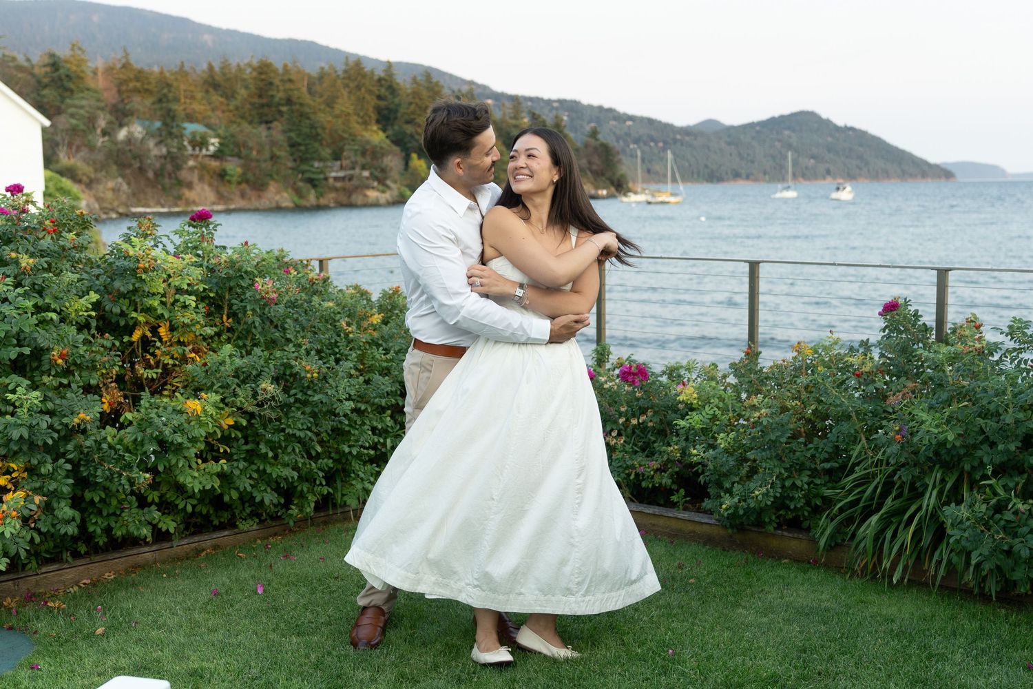 Couple dancing joyfully in garden with scenic waterfront and mountains backdrop on their wedding day.