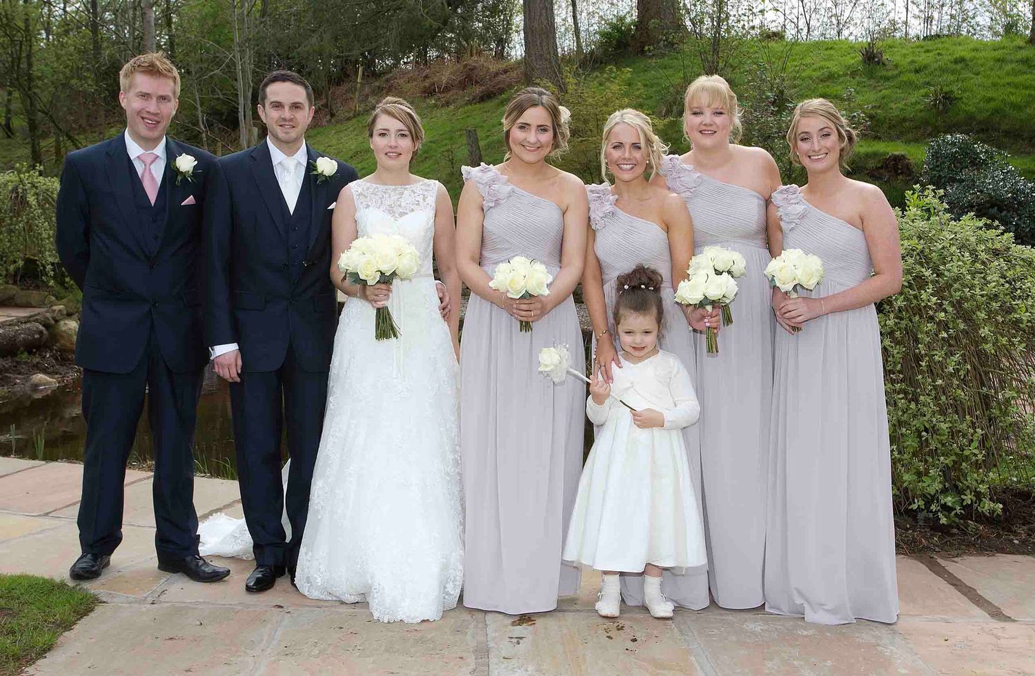Bride and groom posing outdoors with their wedding party, including bridesmaids in pale grey dresses and a flower girl in white.