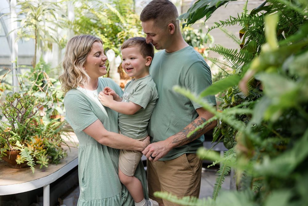 Smiling family of three in matching sage green outfits posing together among lush green plants in a bright greenhouse.
