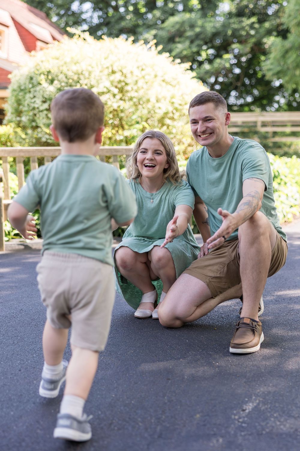 Smiling parents in matching green outfits kneel on a path with open arms as their toddler walks toward them outdoors.