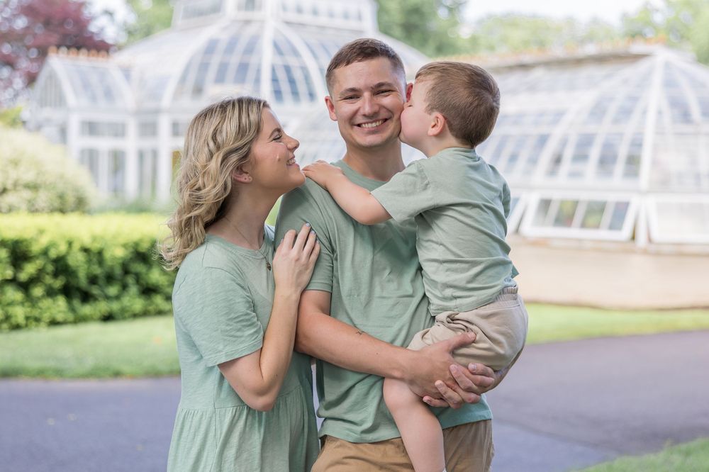 Smiling family in matching sage green outfits embrace outdoors as a young boy kisses his mother's cheek in front of a glass greenhouse.