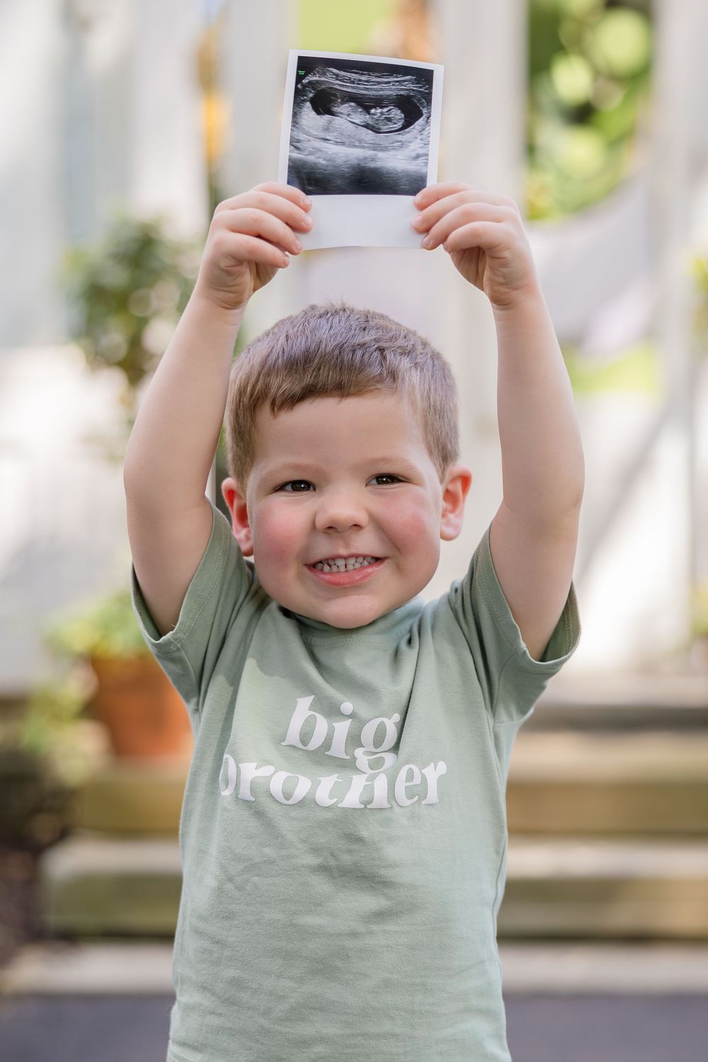 Smiling toddler in a green big brother t-shirt holds an ultrasound photo above his head to announce a new baby sibling.