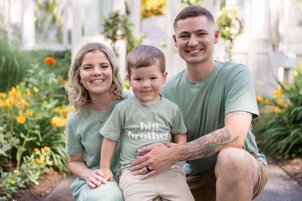 Smiling couple in matching green shirts with their young son in a "big brother" shirt posing among orange flowers in a sunny garden.