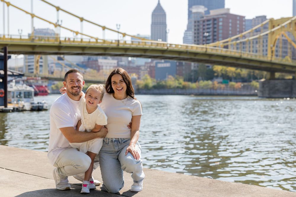 Smiling family of three in white shirts crouching on a riverfront walkway with a yellow suspension bridge and city skyline behind them.