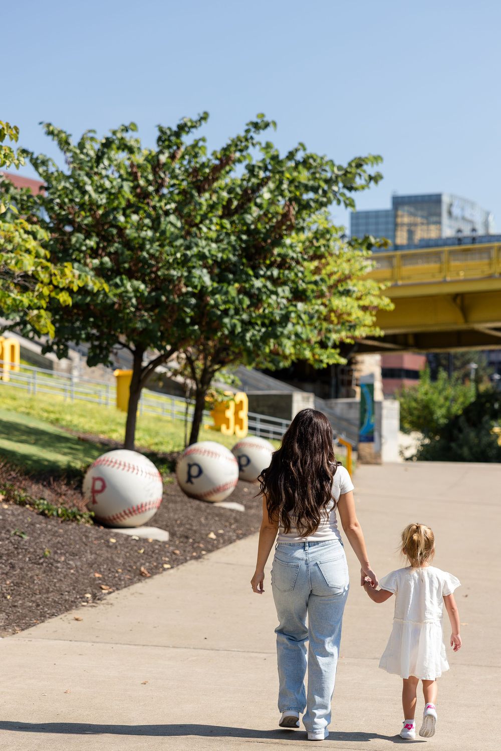A woman and a toddler hold hands while walking along a sunny pathway past giant baseball sculptures and trees near a yellow bridge.