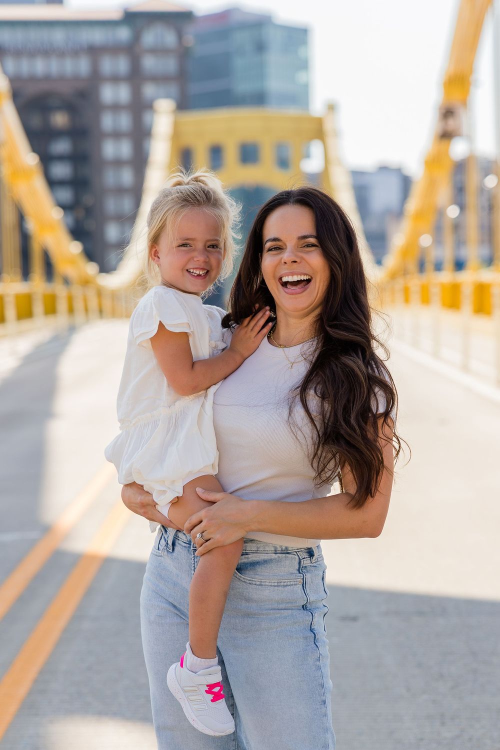 A laughing woman with long dark hair holds a smiling toddler in a white dress on a sunny yellow suspension bridge.