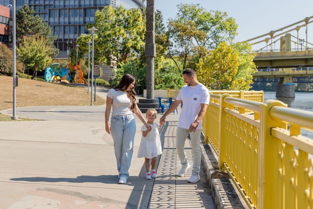 Family of three holding hands and walking with a young girl in a white dress along a sunny bridge walkway with yellow railings.