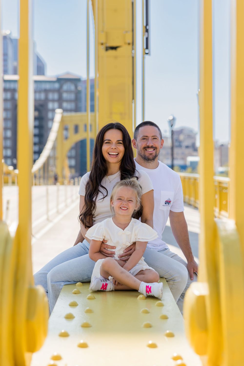 Smiling woman and man sit with a young blonde girl on a bright yellow steel bridge with city buildings in the background.
