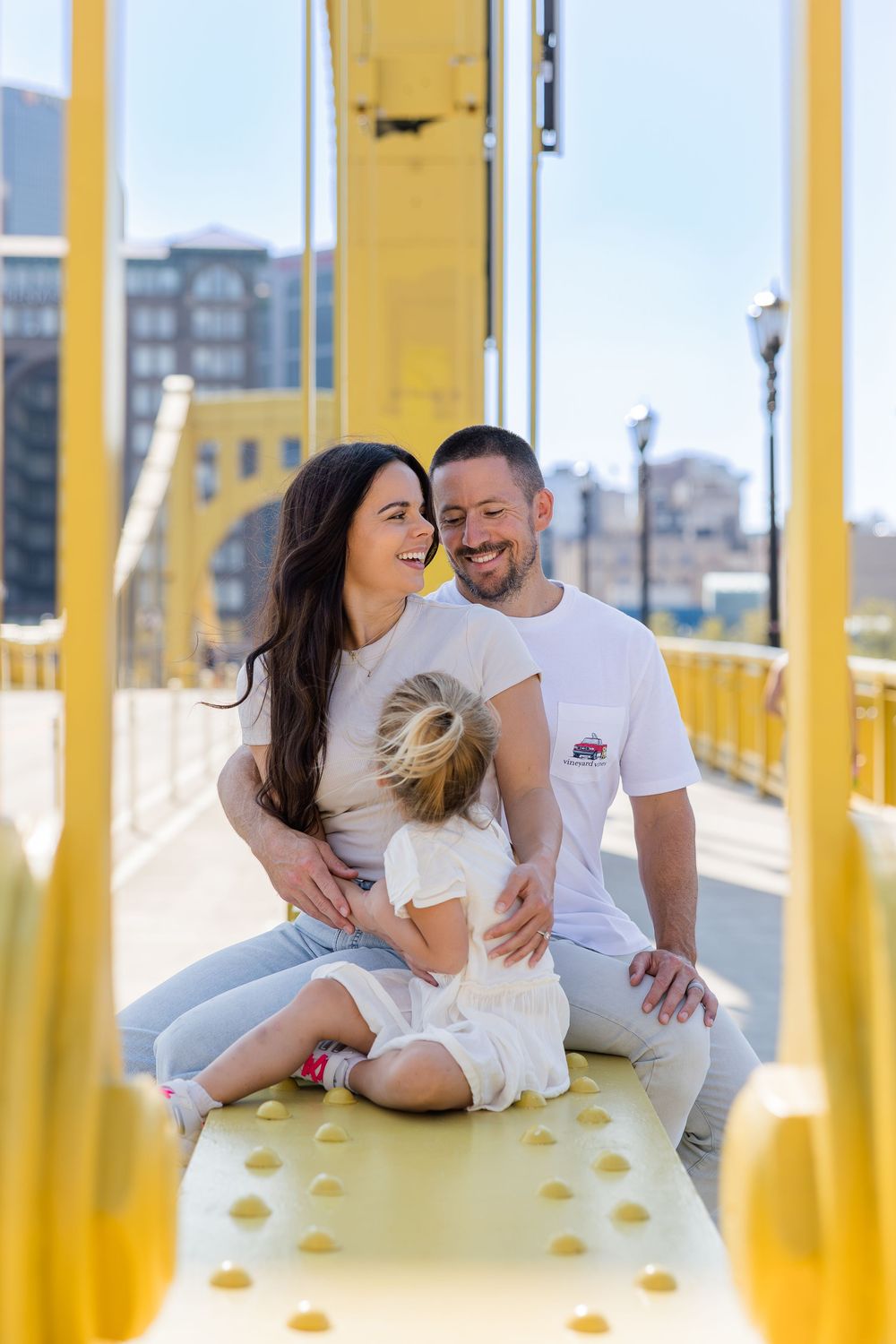 Smiling couple in white shirts sit with their young daughter on a bright yellow steel bridge with city buildings behind them.