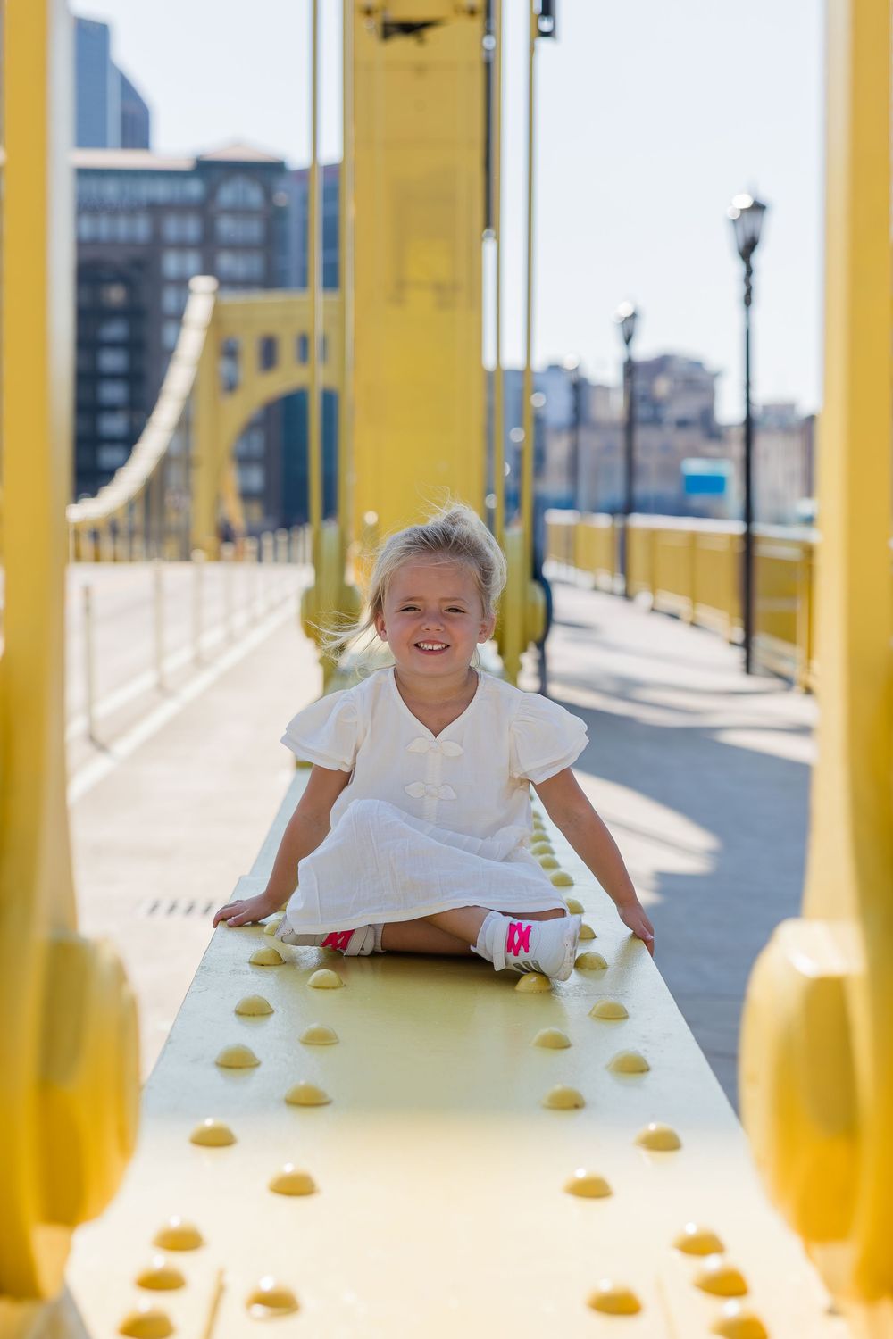 Smiling young girl in a white dress sits on the yellow steel beam of a bridge on a sunny day with city buildings and lampposts behind her.