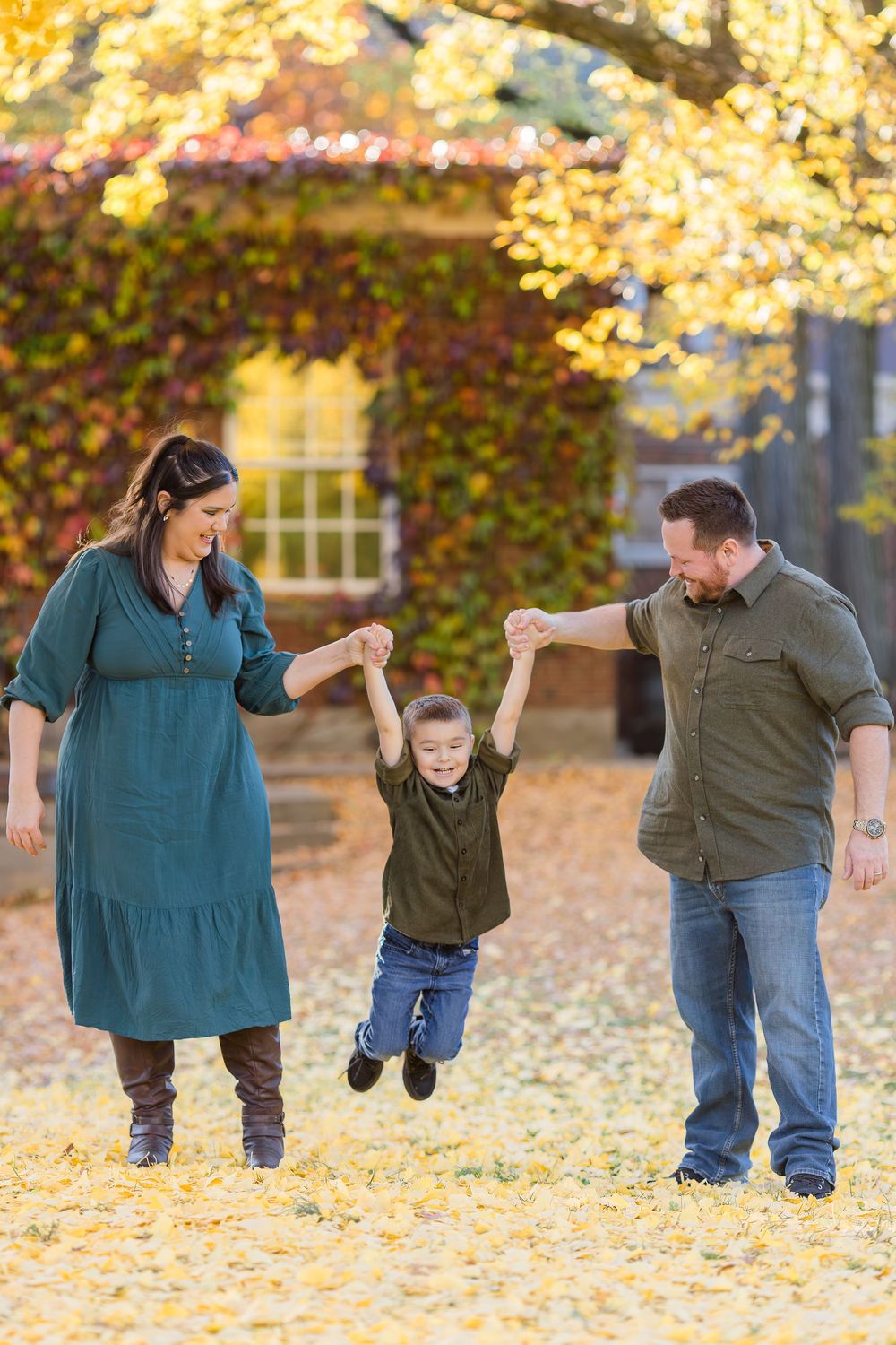 A smiling couple swings their laughing young son between them on a carpet of golden fallen leaves in front of an ivy-covered building.