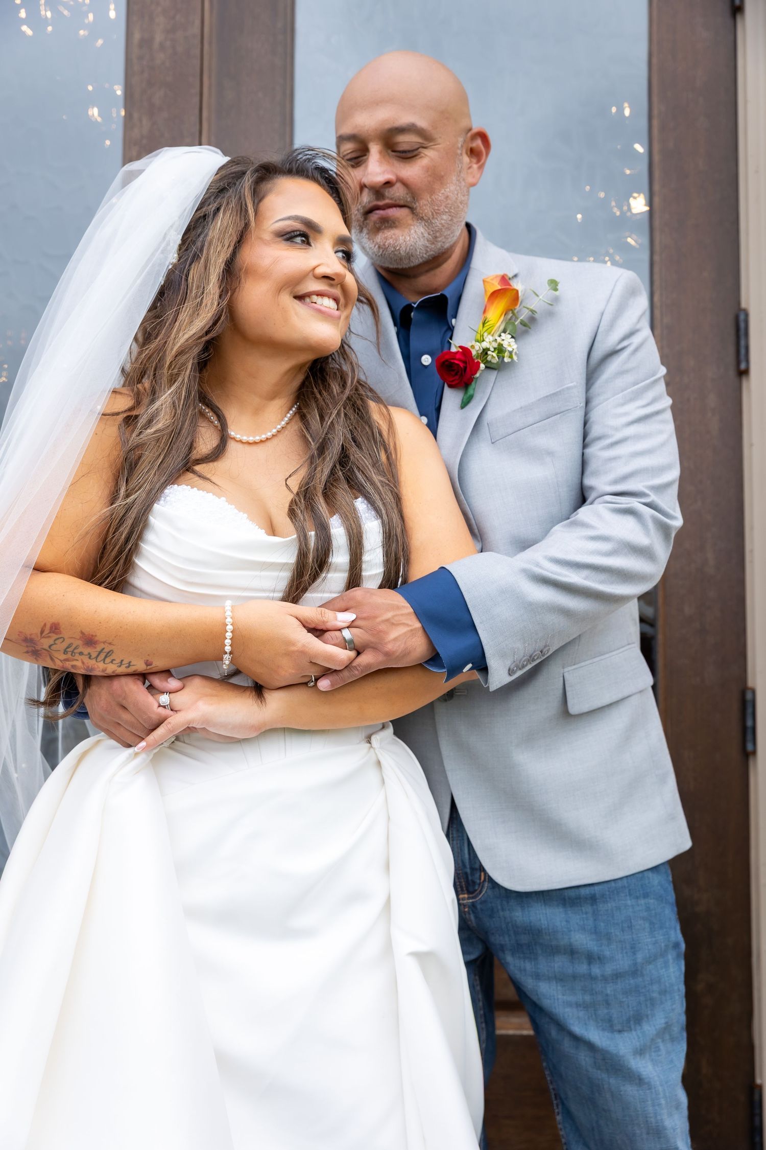 A bride in a white veil kisses her groom in a gray suit outdoors, with colorful red and orange wedding flowers in the foreground.