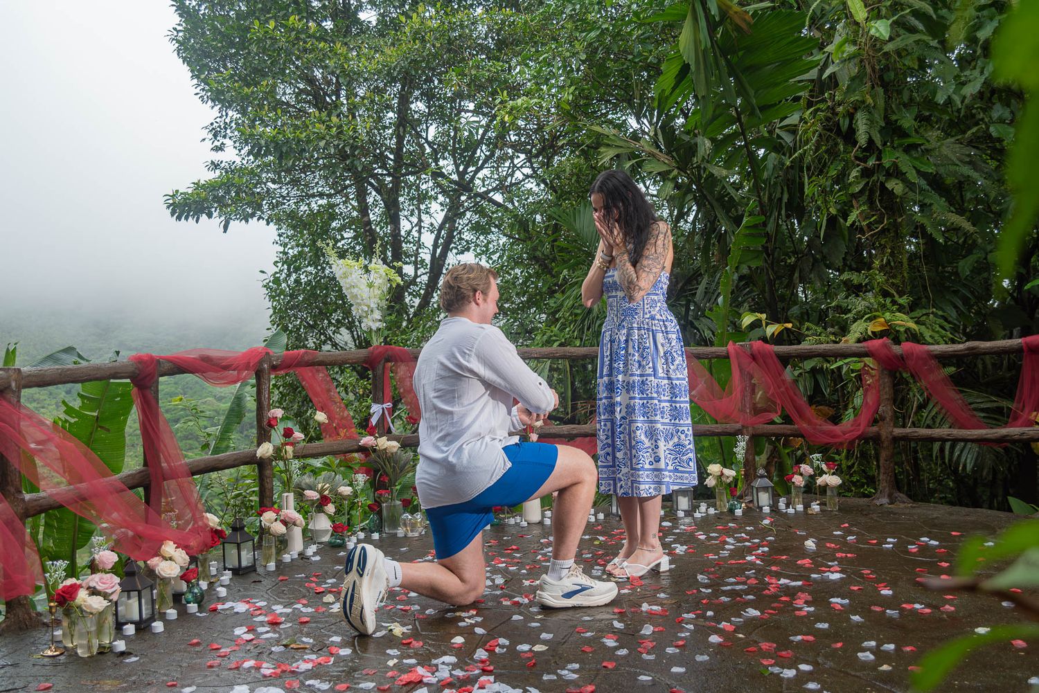 Man kneels on one knee proposing to a woman in a blue dress on a rose petal-covered jungle deck with red draped fabric and candles.