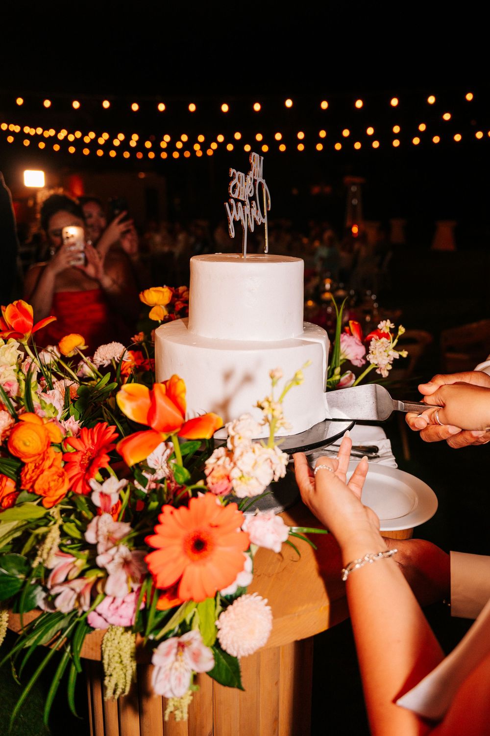 Couple cutting a two-tier white wedding cake with a topper, beside colorful orange and pink floral arrangements under outdoor string lights.