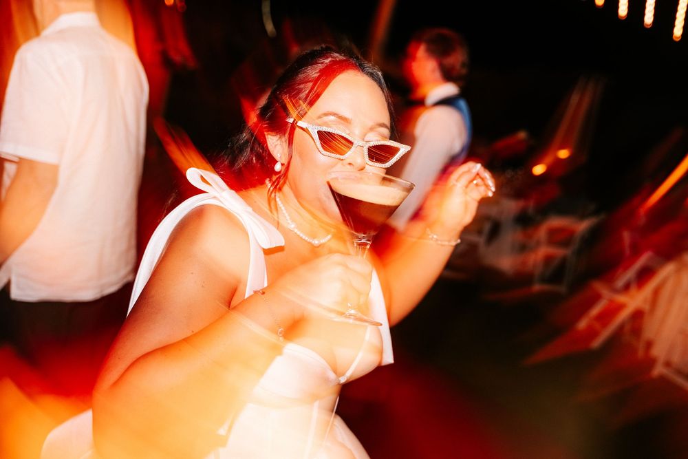 Smiling woman in a white dress and white cat-eye sunglasses holds a drink while dancing at a lively evening party with motion blur.