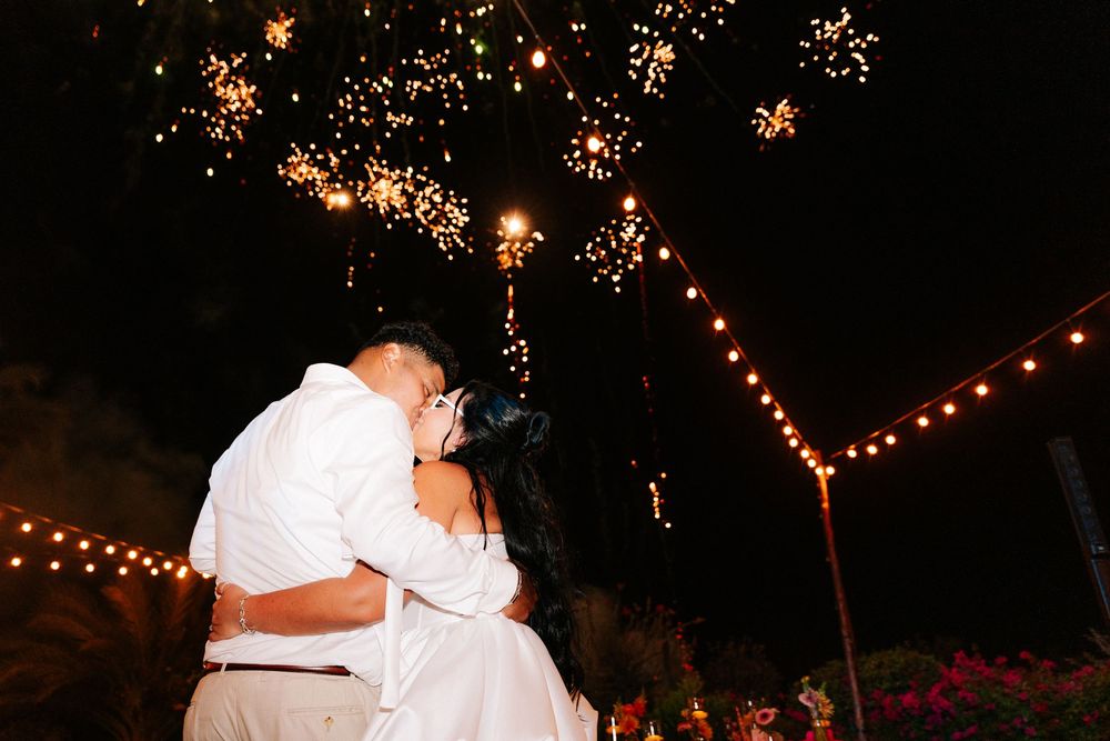 Couple in white embracing and kissing at night under sparkling fireworks and warm string lights at an outdoor wedding celebration.