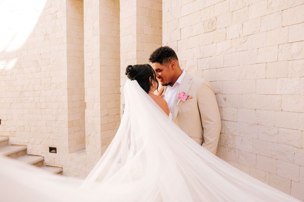 Bride and groom share an intimate moment against a stone wall as her long, flowing wedding veil billows in the breeze.
