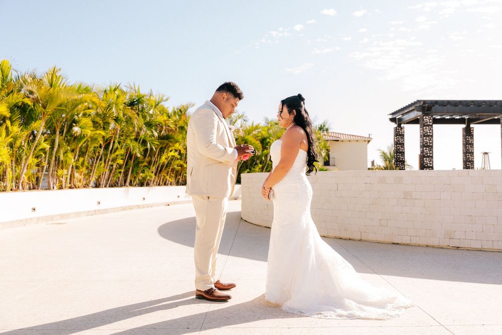 Bride in a flowing white gown and groom in a cream suit share a private moment on a sunny terrace with palm trees and a pergola.