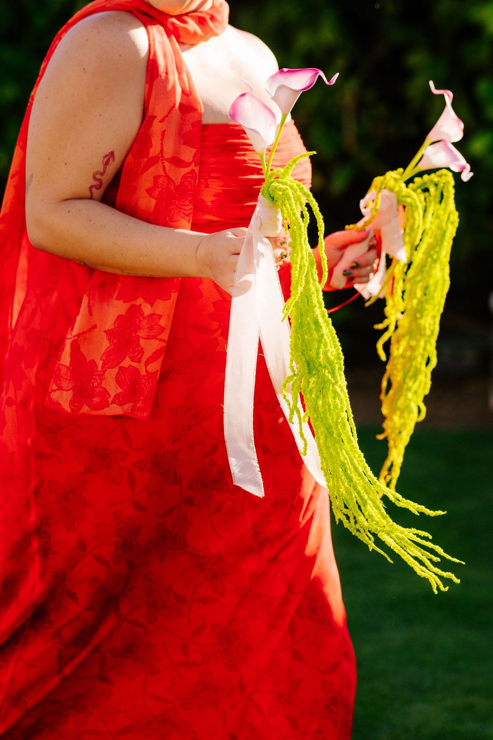 Woman in a red lace dress holds a bouquet of pink calla lilies and trailing green amaranthus tied with a white ribbon outdoors.