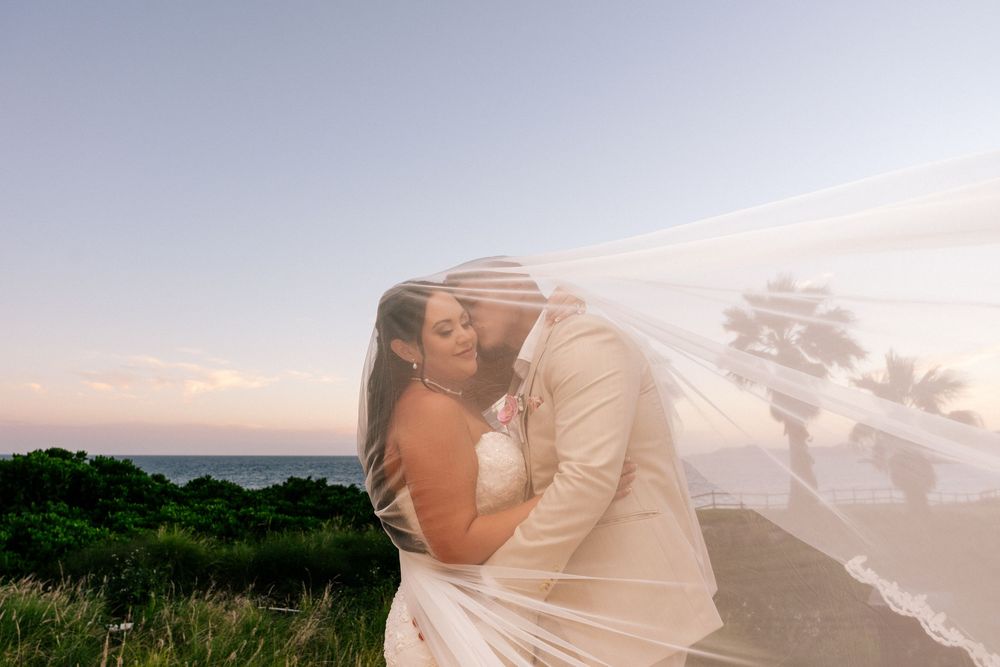 Bride and groom embrace under a sheer veil at sunset, with palm trees and the ocean glowing in the background.