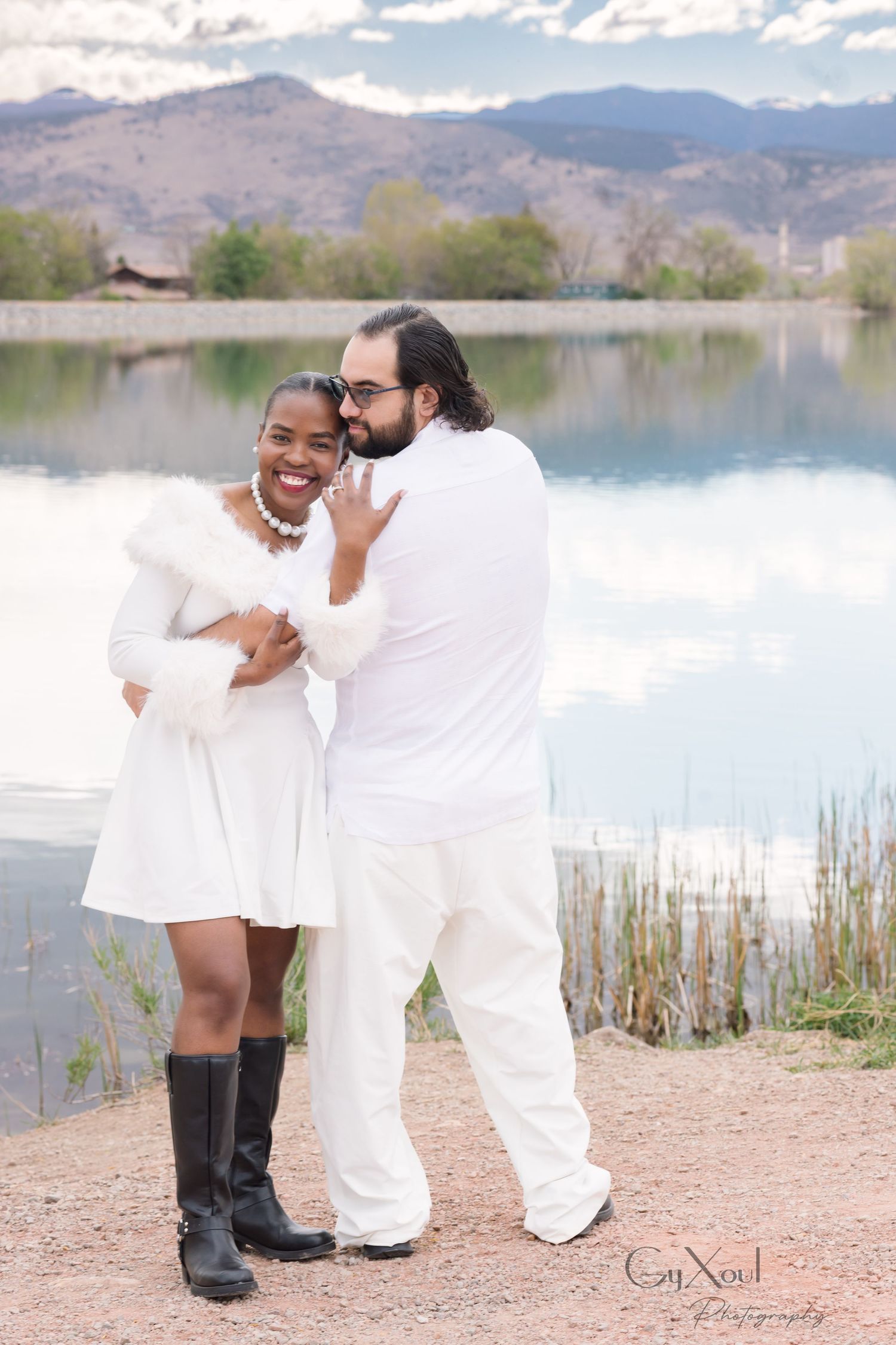 Smiling couple in white outfits embrace on a lakeside shore with mountains and reeds in the background during an engagement photo session.