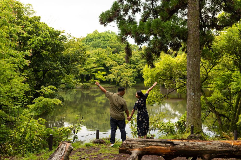 Couple holding hands with arms raised stand beside a calm pond surrounded by lush green trees in a peaceful garden.