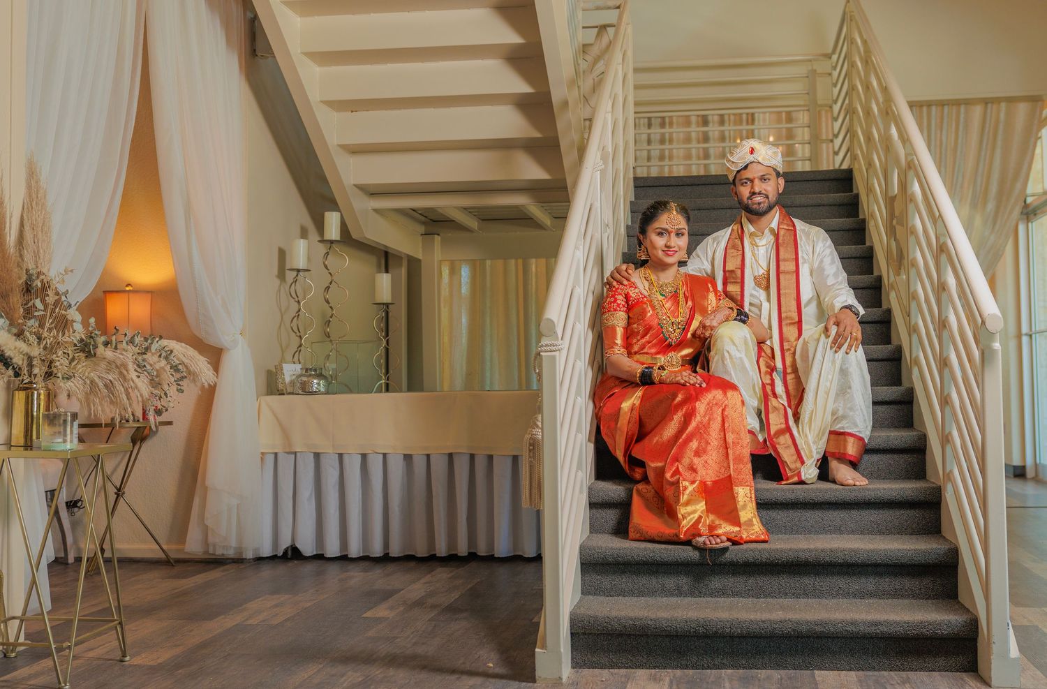 Indian couple in traditional wedding attire sitting on stairs, bride in red saree with gold jewelry, groom in white sherwani.