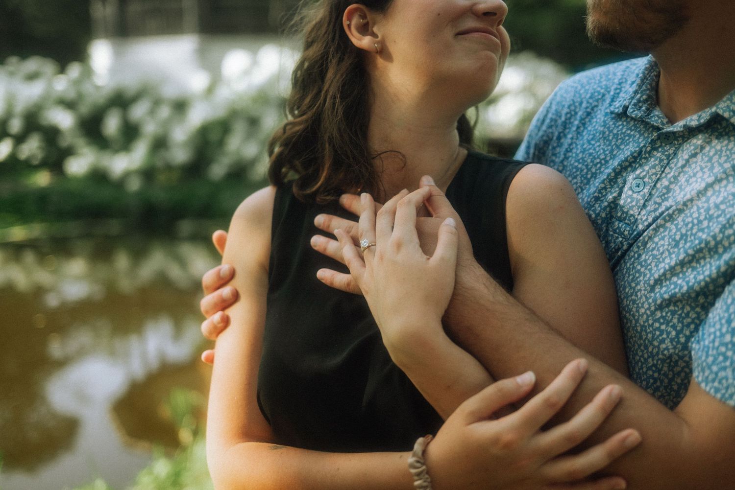Smiling woman in a black dress leans into a man's embrace, showing her ring while standing beside a tranquil pond with greenery. Vondelpark