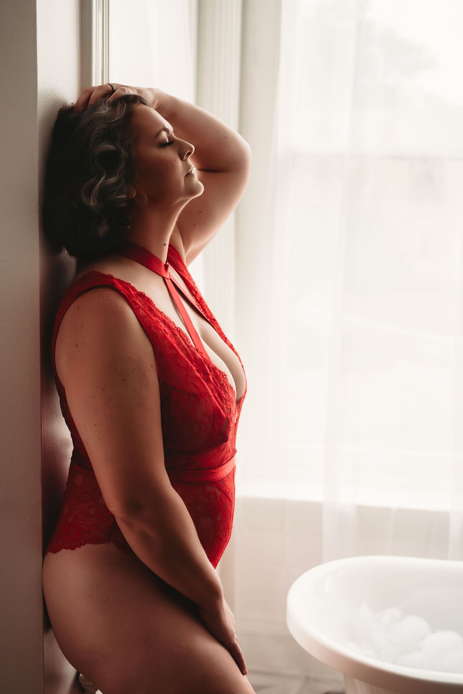 Woman in a red lace bodysuit leans against a wall with her eyes closed beside a bubble bath in a bright window-lit boudoir photo session.
