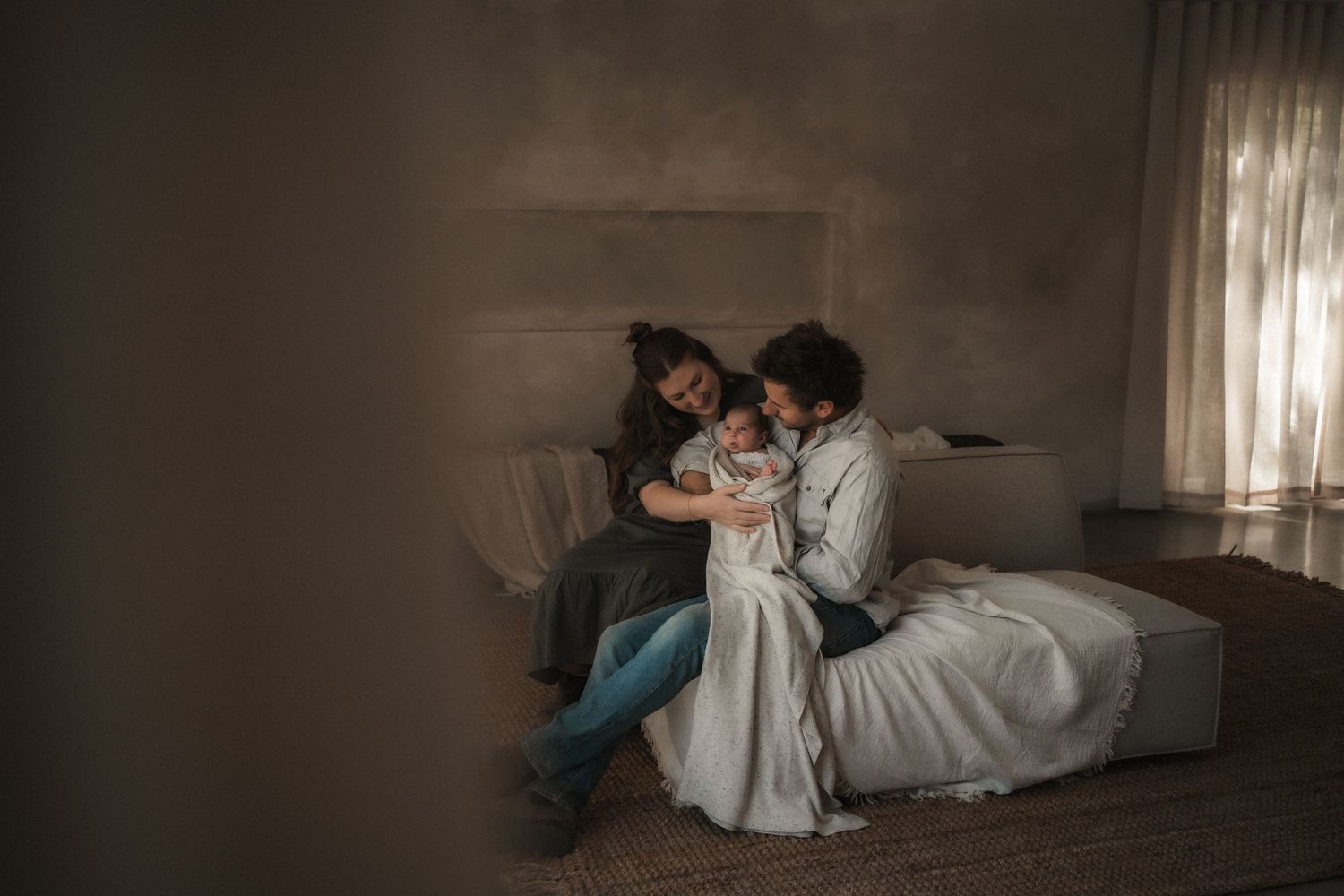 Couple sitting together on a bed in a softly lit room, gently cuddling their newborn baby wrapped in a white blanket.