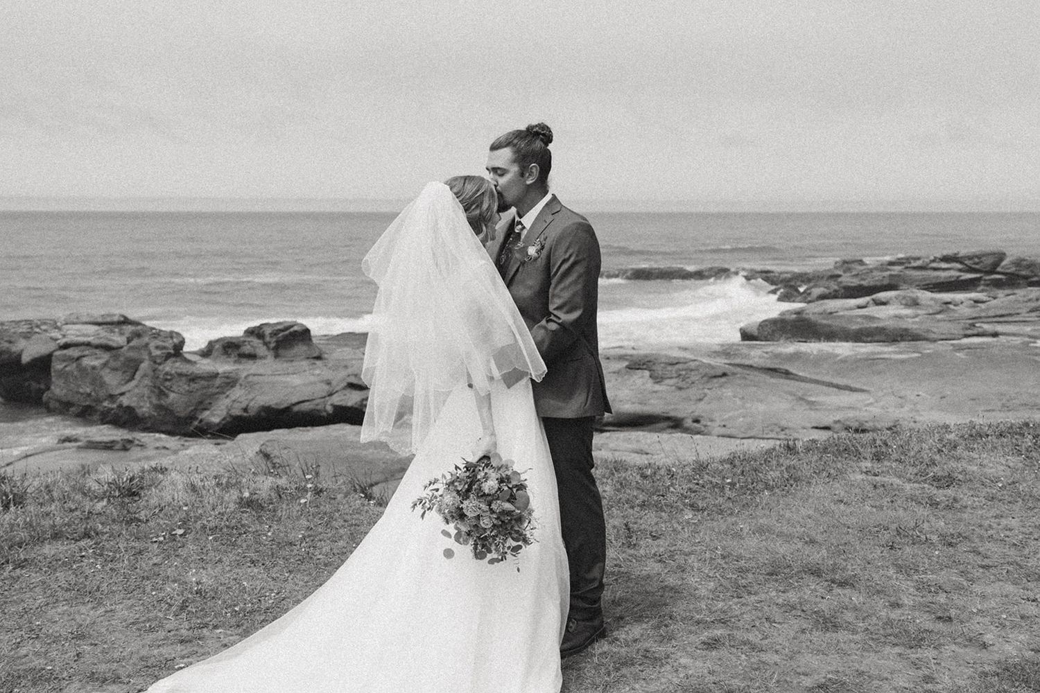 Small outdoor wedding ceremony on a coastal cliff with ocean views, guests gathered around bride and groom under cloudy skies.
