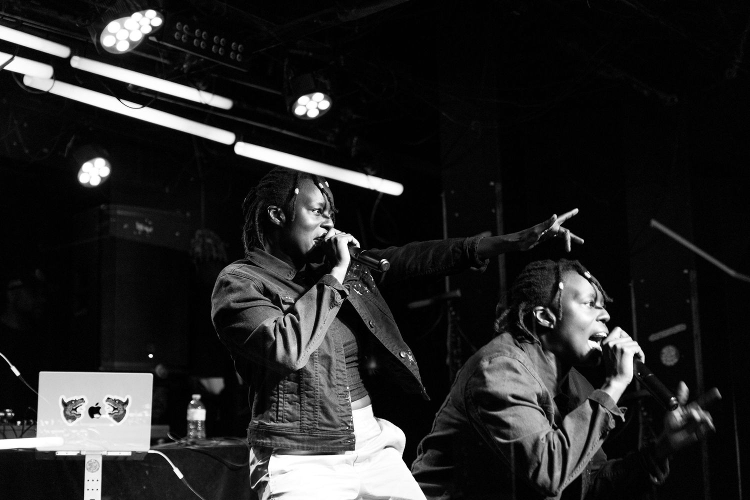 Two Black male performers singing on stage under bright lights in a black and white concert photo.