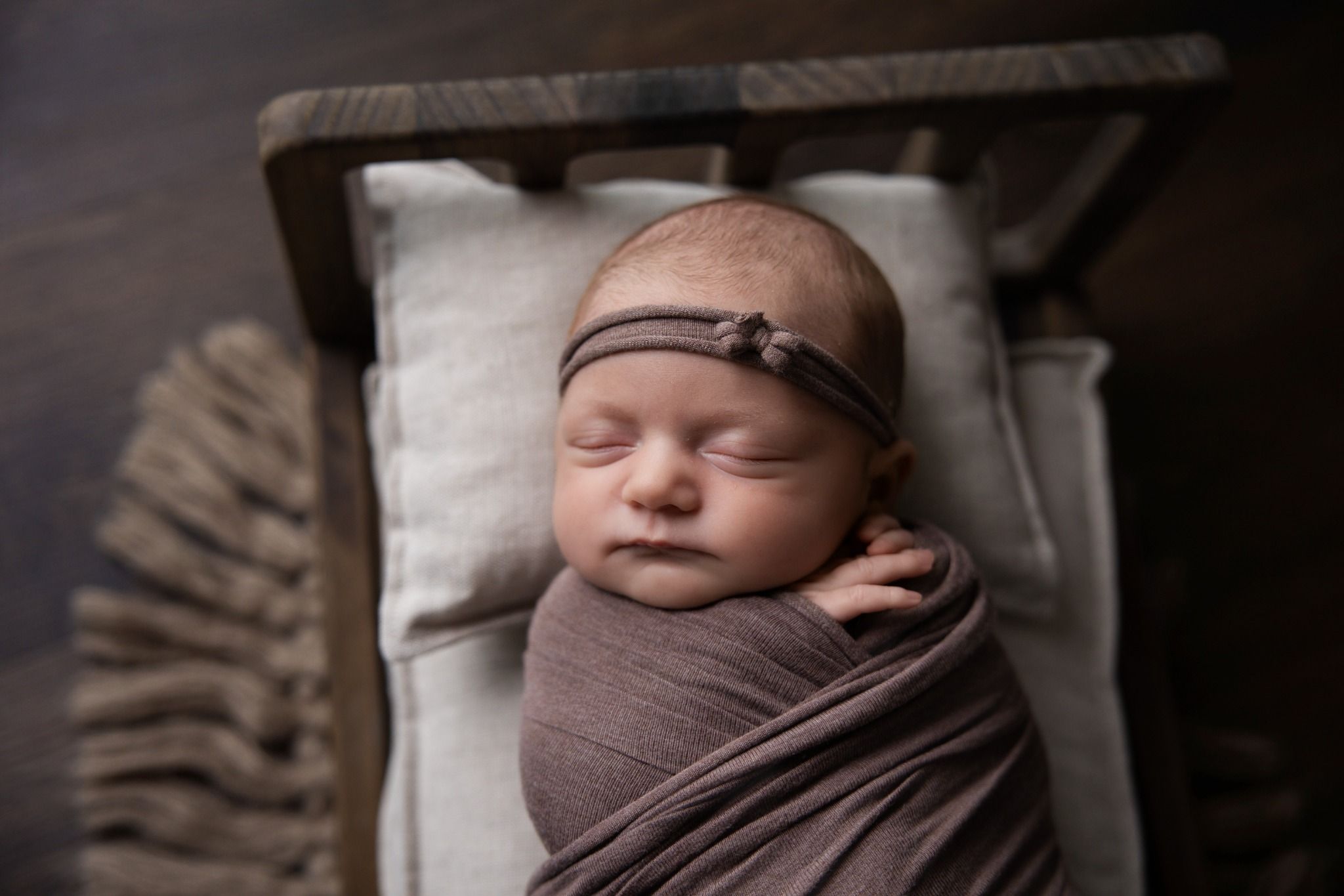 Sleeping newborn wearing a knotted headband and wrapped in mauve on a small wooden bed