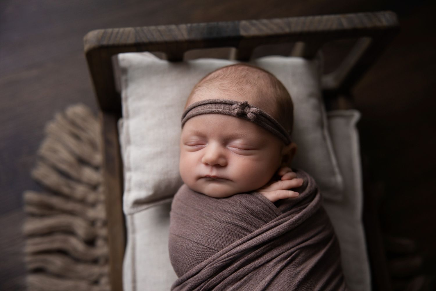 Sleeping newborn baby wrapped in a mauve swaddle with a matching headband, resting on a small wooden bed with a cream pillow.