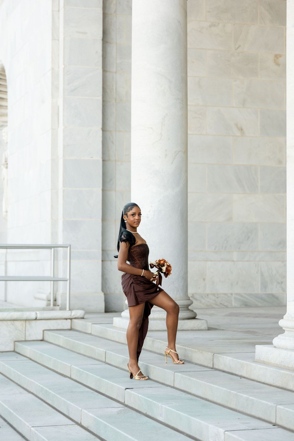 Woman in a brown dress and heeled sandals poses with a bouquet on marble steps beside large neoclassical columns.