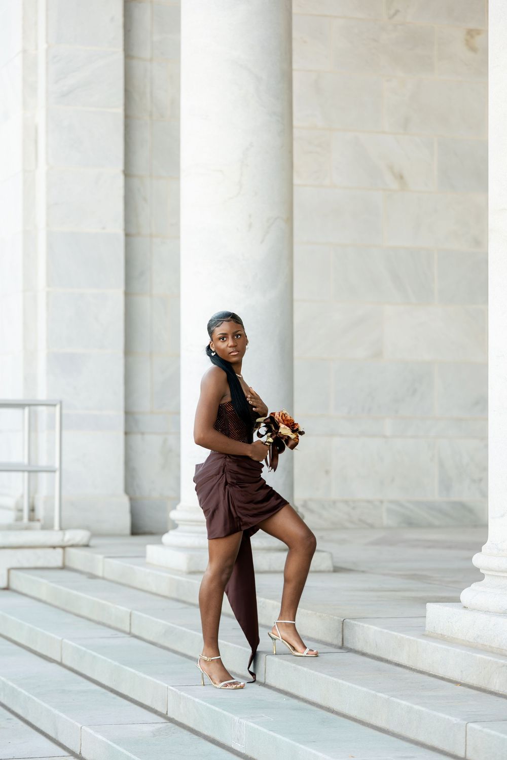 Woman in a burgundy wrap dress and strappy heels holds a floral bouquet while posing on white marble steps beside tall columns.