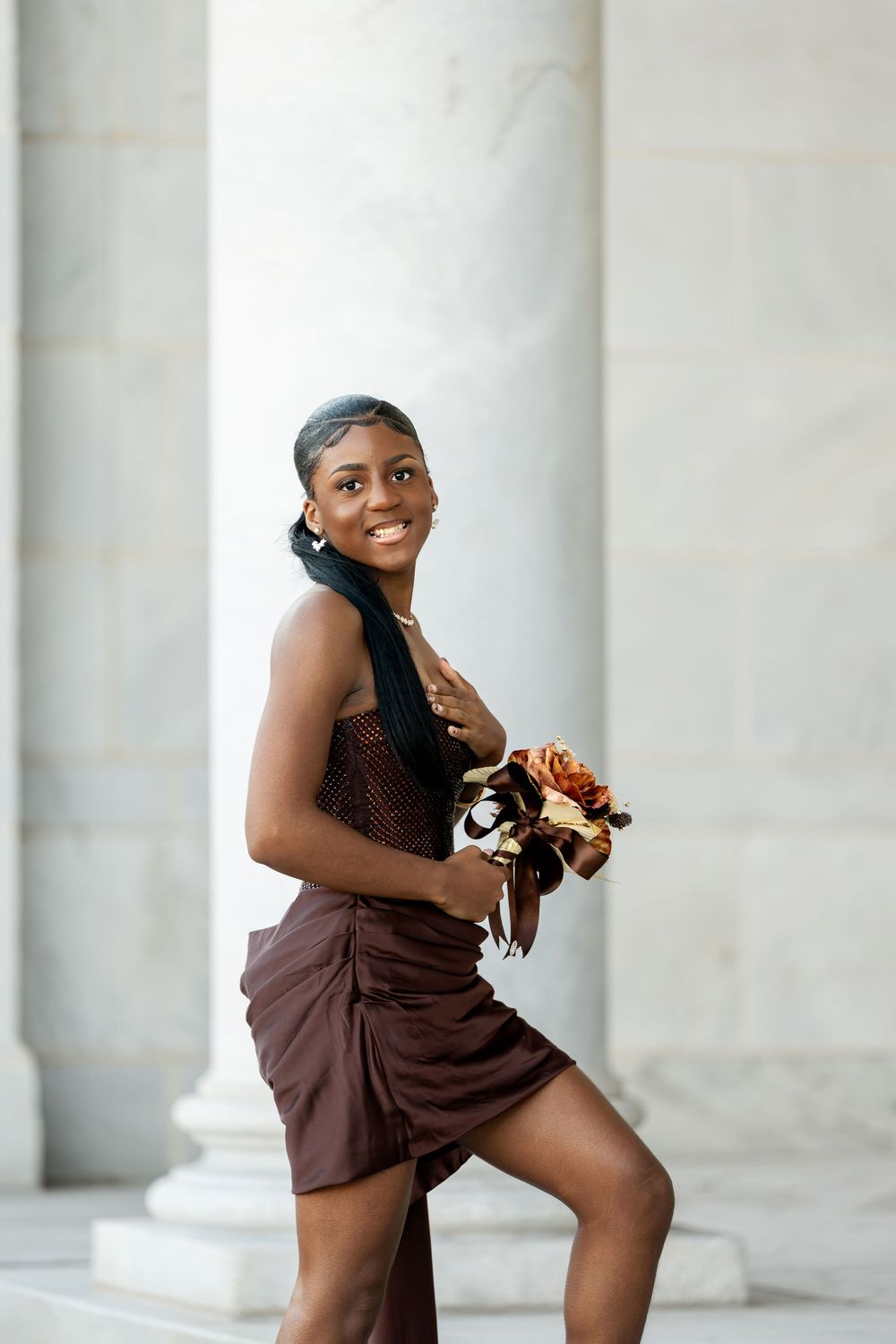 A smiling woman in a brown strapless dress holds a copper bouquet while posing against tall white marble columns.