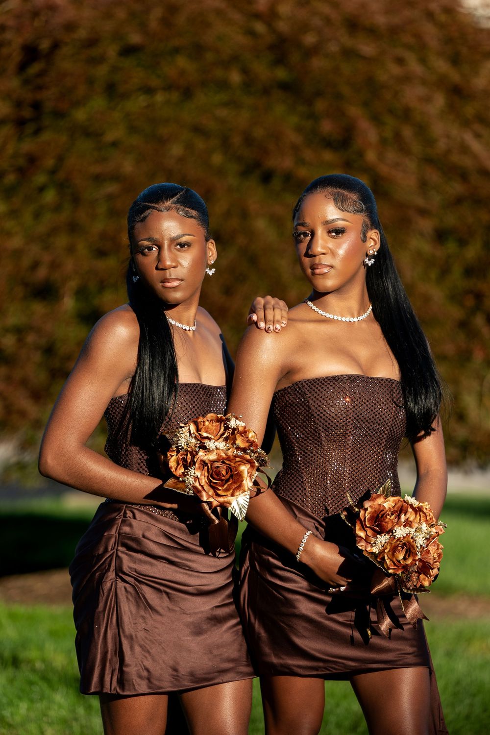 Two bridesmaids in brown satin dresses and pearl necklaces hold copper rose bouquets outdoors in front of autumn foliage.