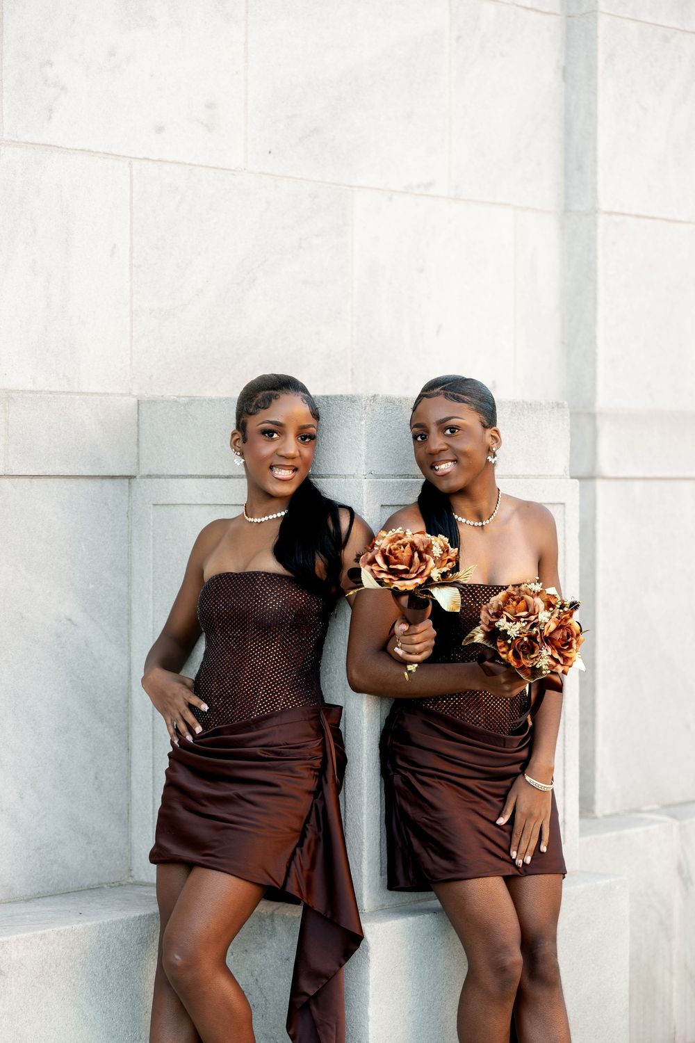 Two smiling bridesmaids in brown satin dresses pose against a white marble wall while holding copper rose wedding bouquets.