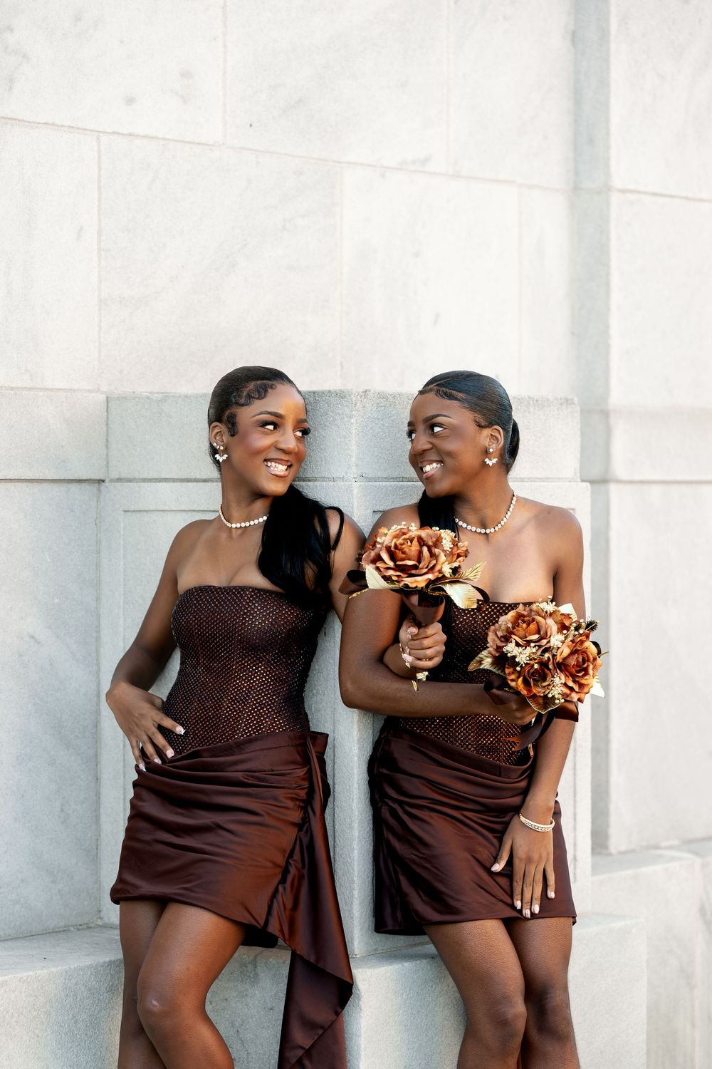 Two smiling bridesmaids in brown satin dresses and pearl necklaces hold copper rose bouquets while leaning against a white marble wall.