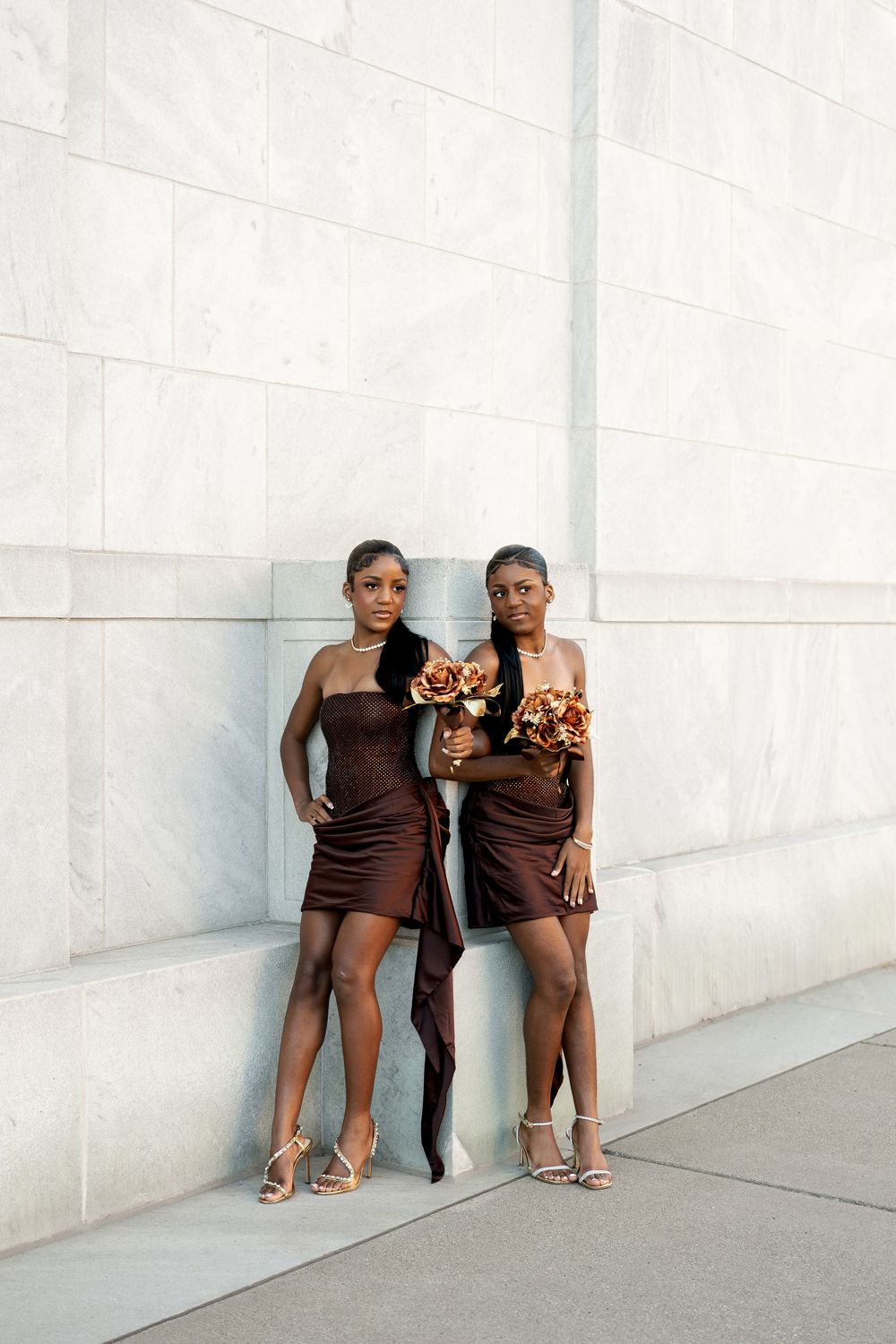 Two bridesmaids in chocolate brown dresses and strappy heels pose with copper flower bouquets against a white stone building wall.