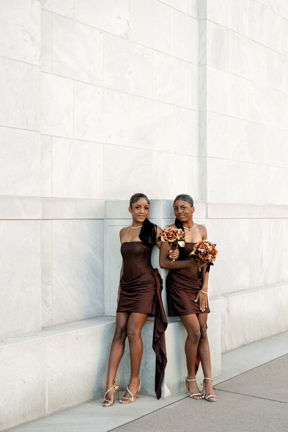 Two women in brown satin dresses and heels pose with autumn bouquets against a white marble wall.