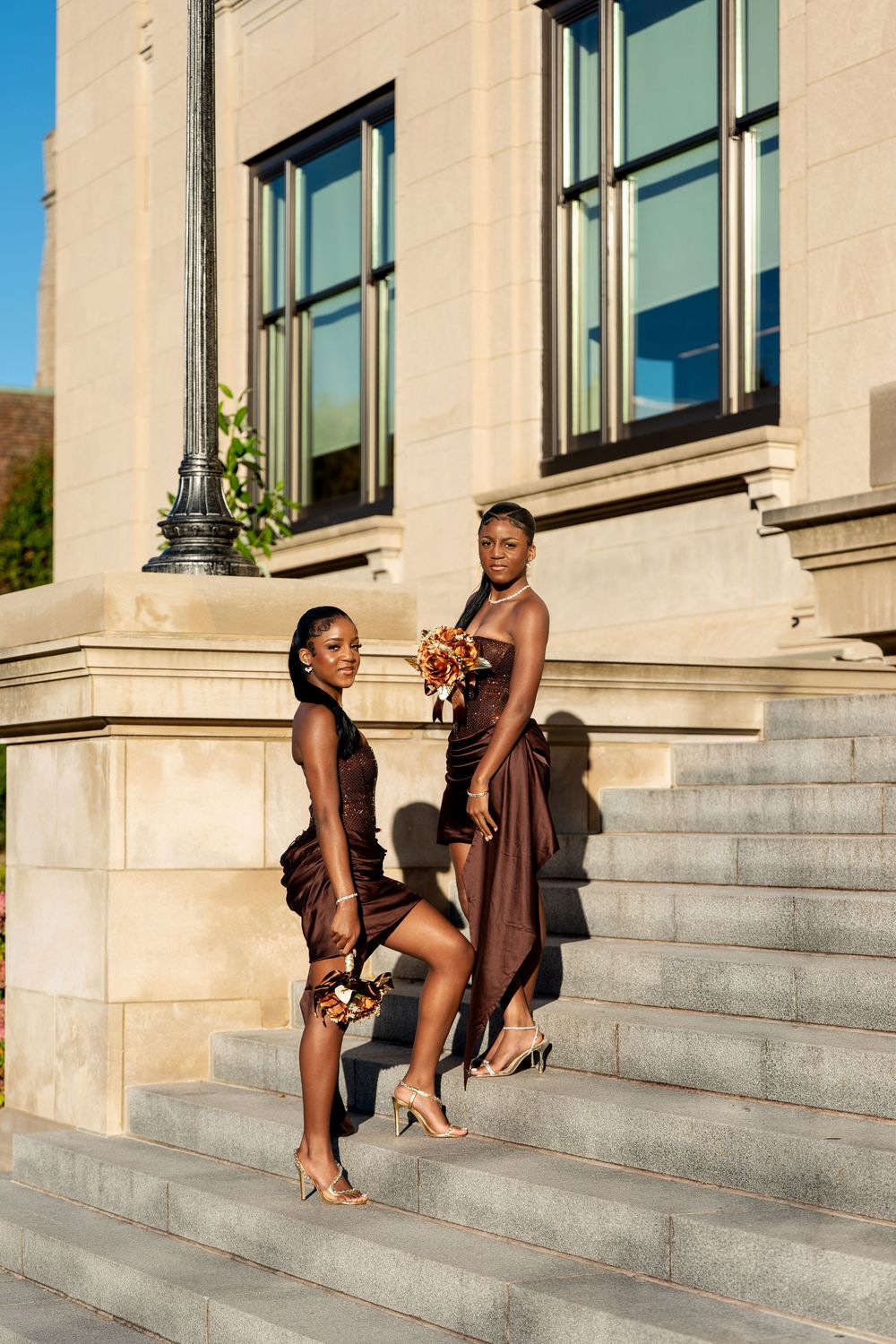 Two women in chocolate brown dresses and strappy heels stand on granite steps holding autumn-toned bouquets.