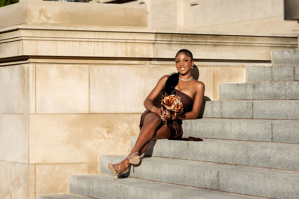 Woman in a brown satin dress sits on gray stone steps holding an orange autumn bouquet, smiling in warm sunlight.