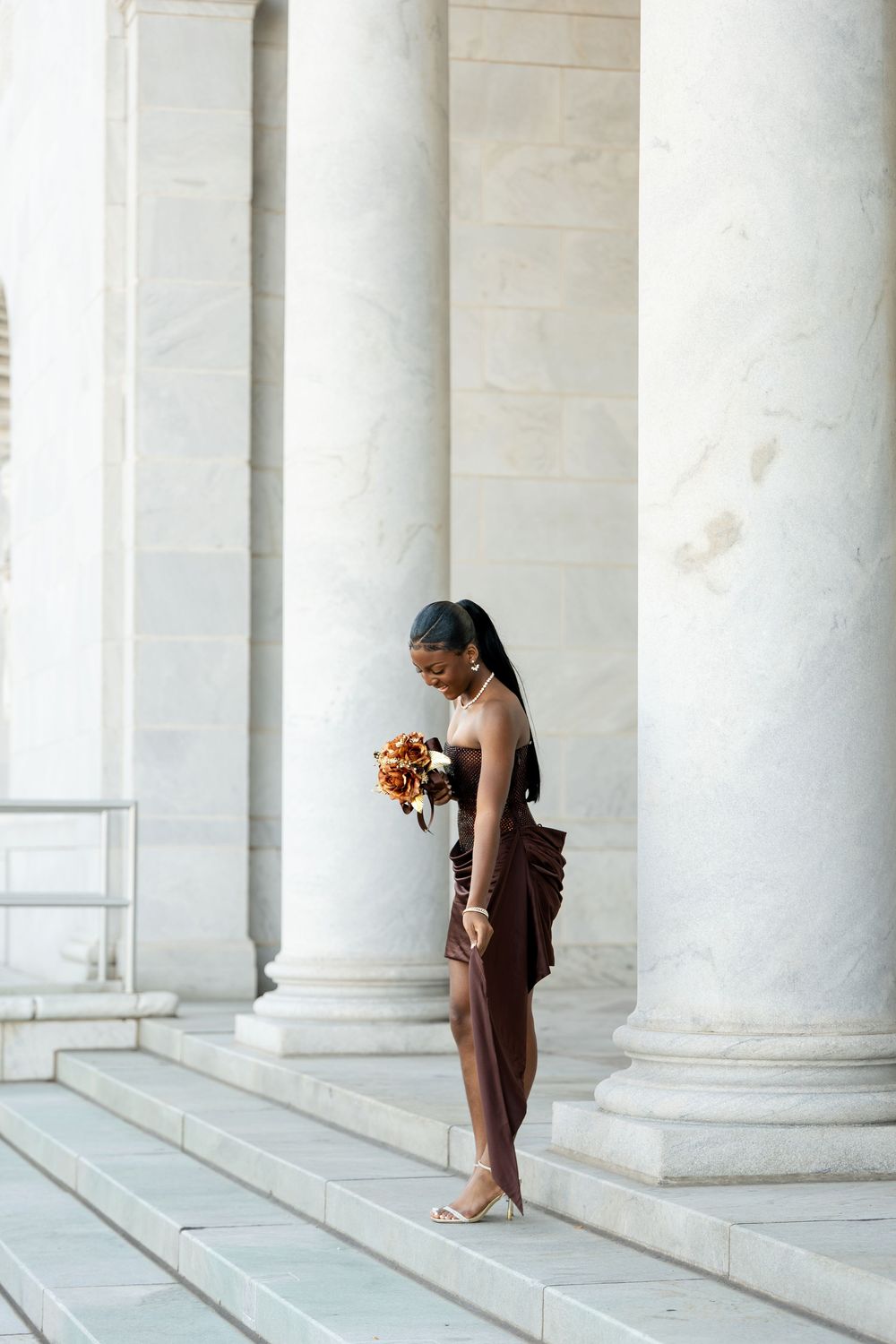 Woman in a brown strapless dress holding an orange bouquet walks down white marble steps between tall stone columns.